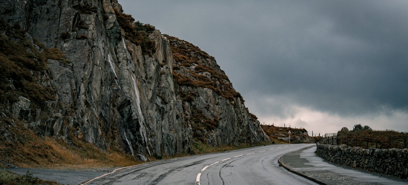 A wet mountain road curving away beneath a rock face under heavy cloud